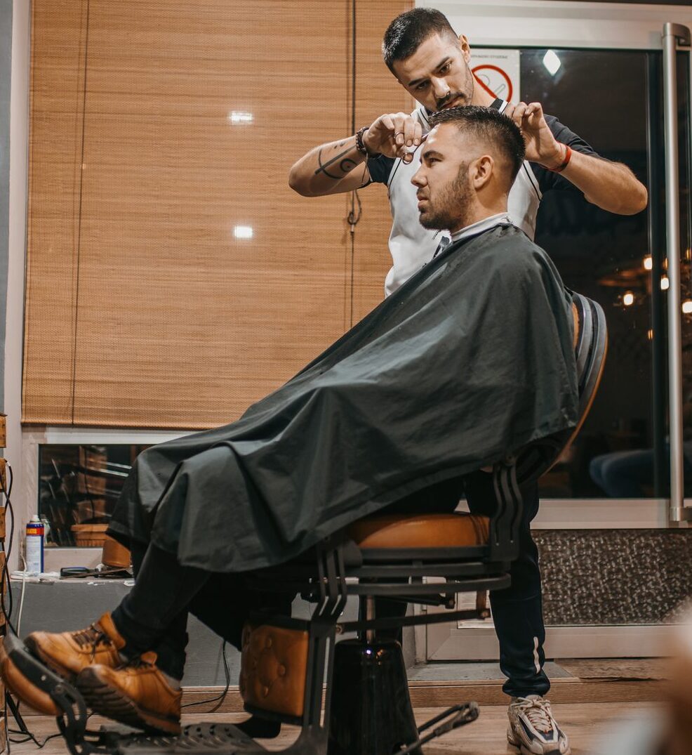 Young handsome male client sitting in the chair in the barber shop while male handsome barber cutting his hair